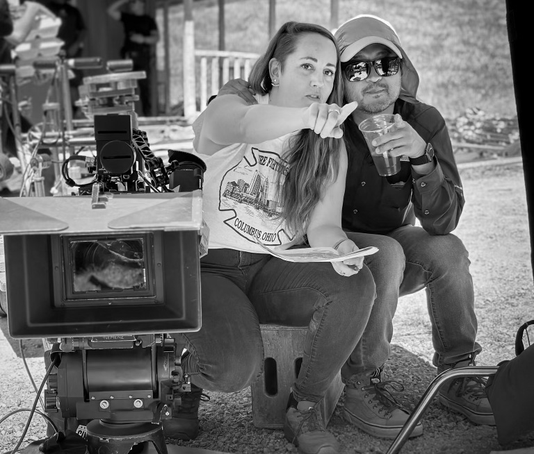 Grayscale photo of a camera on a film set, with Amanda sitting on an apple box next to the camera with a coworker and pointing out to the set
